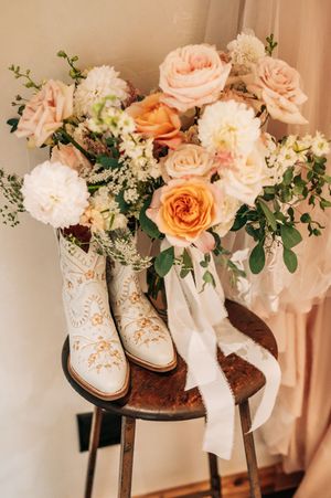 White embroidered cowboy boots displayed on rustic stool with lush peach and white rose bridal bouquet.