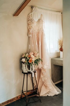 Blush lace long-sleeve wedding dress hanging near window with bridal bouquet and white cowboy boots on stool.