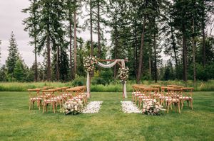 Outdoor wedding ceremony setup with wooden arch, cross-back chairs, and tall pine trees in background.