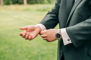 Man in dark suit adjusting cufflinks while wearing a silver watch, standing outdoors on green grass.