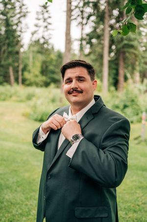 Groom in dark green suit adjusting boutonniere outdoors with tall trees and lush greenery in background.