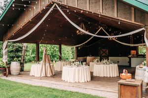 Rustic outdoor wedding reception venue with string lights, draped fabric ceiling, round tables and wooden barrels.