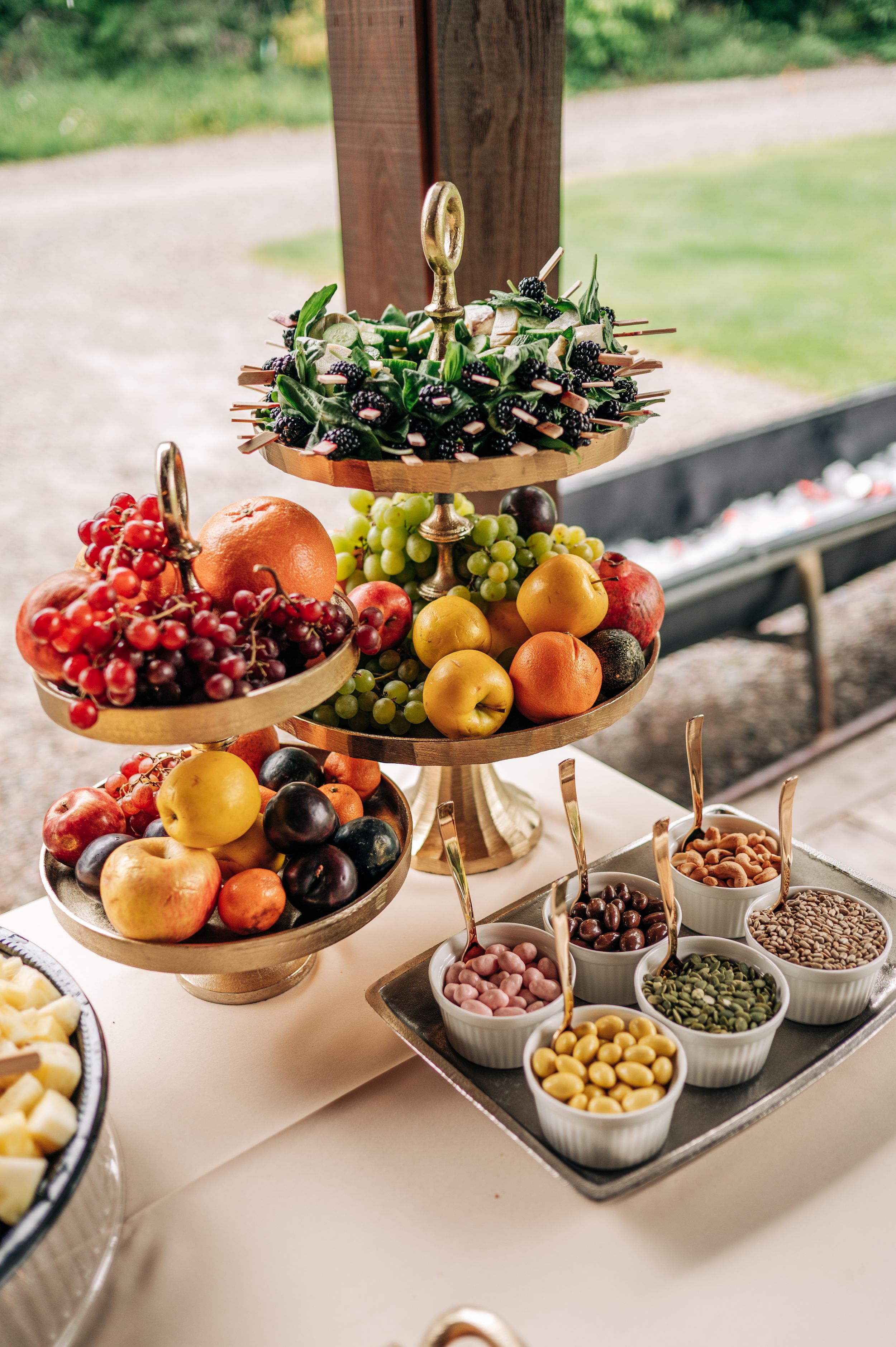 Elegant three-tier gold stand with fresh fruit display and assorted nuts in small bowls at wedding reception.