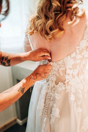Tattooed hands fasten delicate buttons on the back of a bride's floral lace wedding gown.