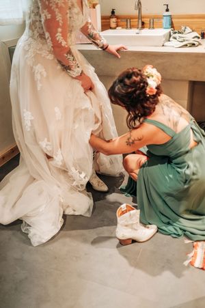 Bridesmaid in green dress helps bride put on white cowboy boots in bathroom before wedding ceremony.