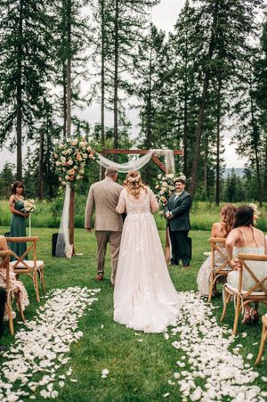 Bride walking down petal-lined aisle toward floral arch at an elegant outdoor forest wedding ceremony.