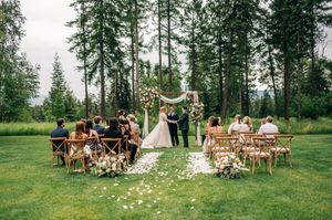 Couple exchanging vows under floral arch surrounded by guests at a scenic outdoor forest wedding.