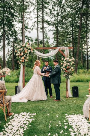 Bride and groom exchange vows under a wooden arch adorned with peach and white flowers in a forest setting.
