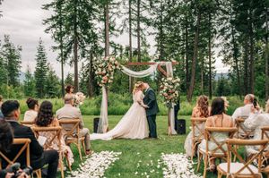 Outdoor wedding ceremony with couple at floral arch altar, seated guests, and tall pine trees in background.
