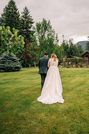Bride in lace gown and groom in dark suit walk hand-in-hand across a lush green lawn.