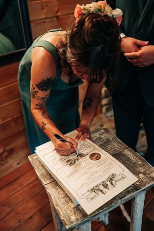Woman in teal bridesmaid dress with floral crown signs official wedding certificate at rustic wooden table.
