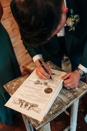 Groom signing wedding marriage certificate document with pen at rustic wooden table during ceremony.