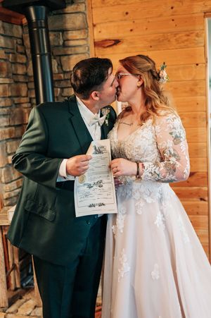 Newlyweds share a kiss while holding their marriage certificate inside a rustic log cabin.