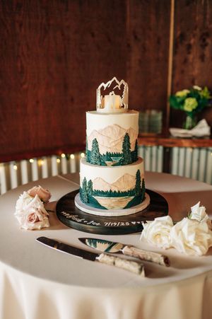 Three-tier mountain themed wedding cake with painted pine trees and peaks displayed on a decorated cake stand.
