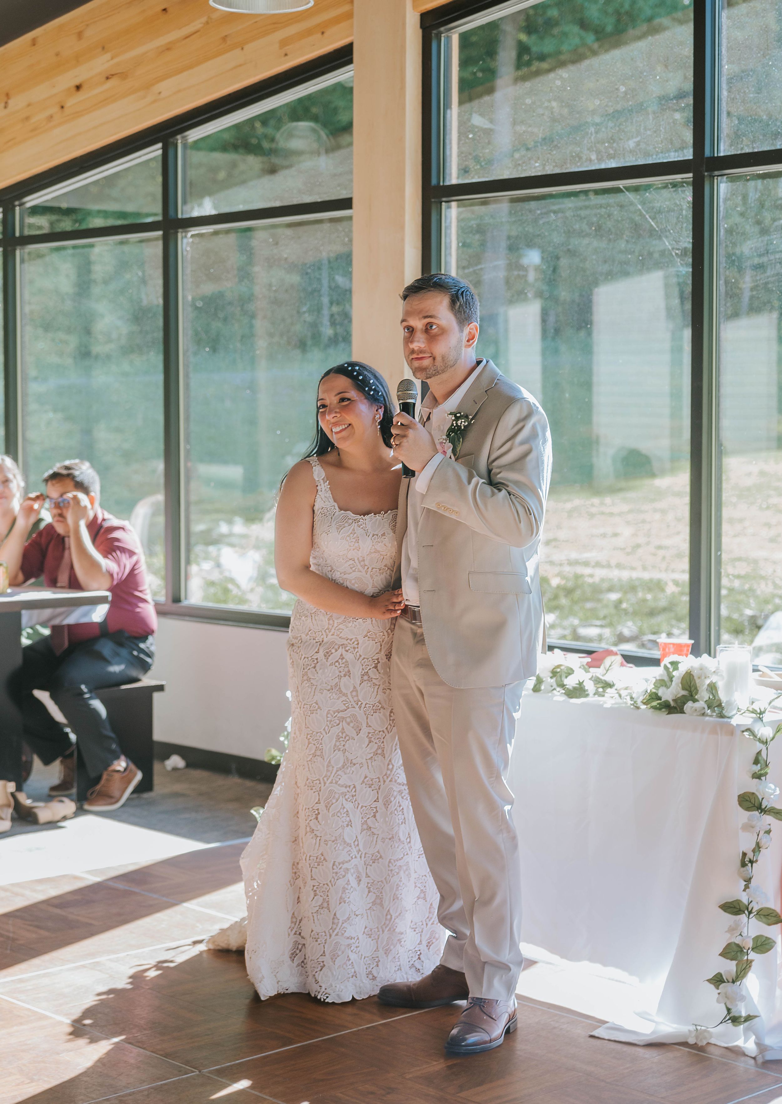Bride and groom sharing a microphone during wedding reception speech in a modern venue with large windows.
