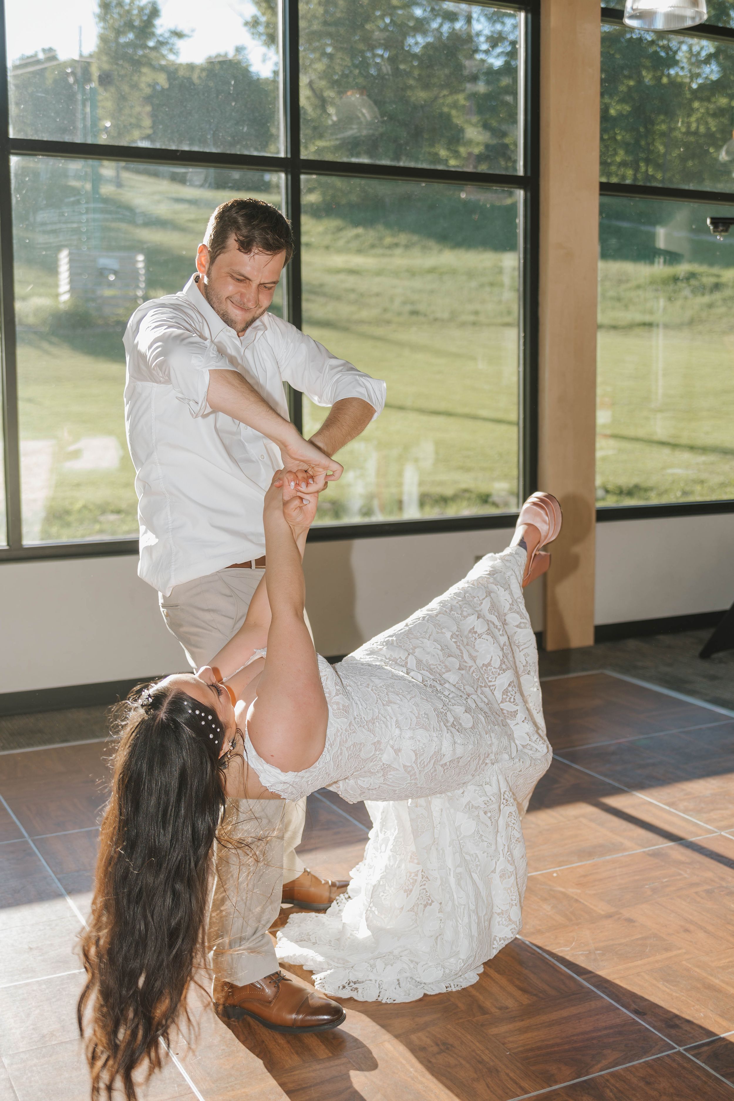 Bride and groom share a joyful first dance, with groom dipping bride in a sunlit wedding venue.