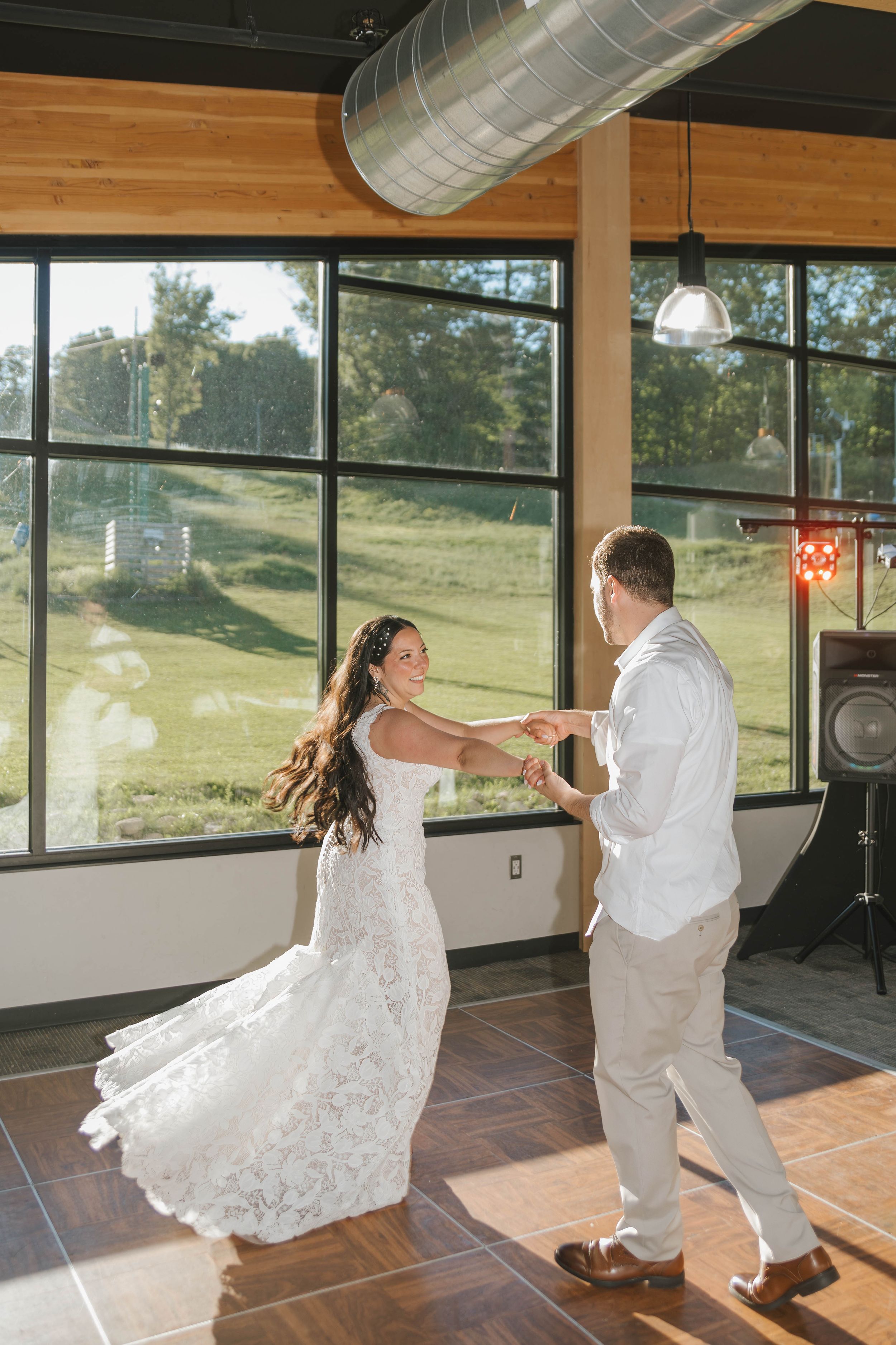 Bride and groom share first dance in sunlit venue with large windows overlooking green landscape on their wedding day.
