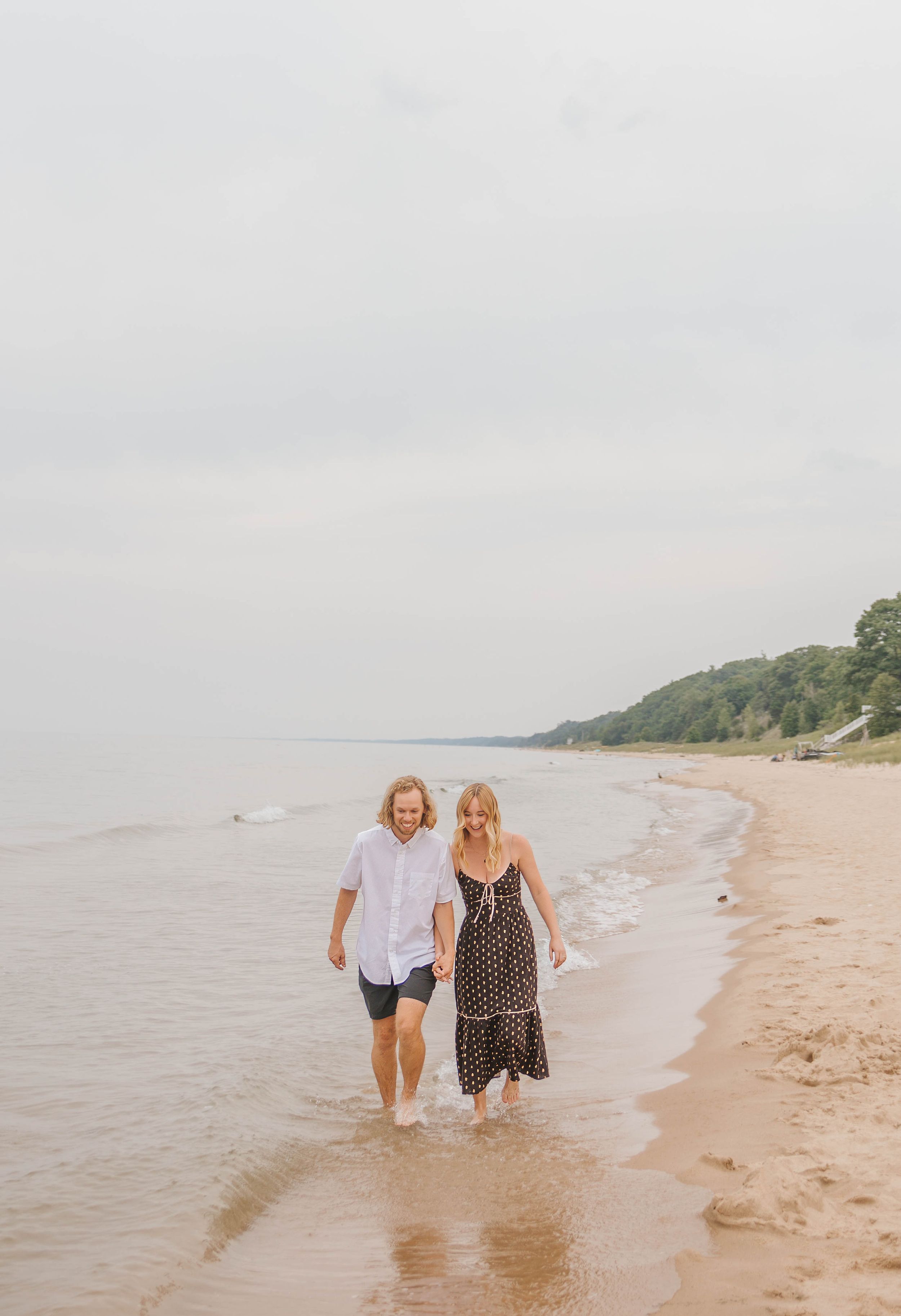 A couple walks hand-in-hand along a sandy beach shoreline, the woman in a floral dress and man in a white shirt.