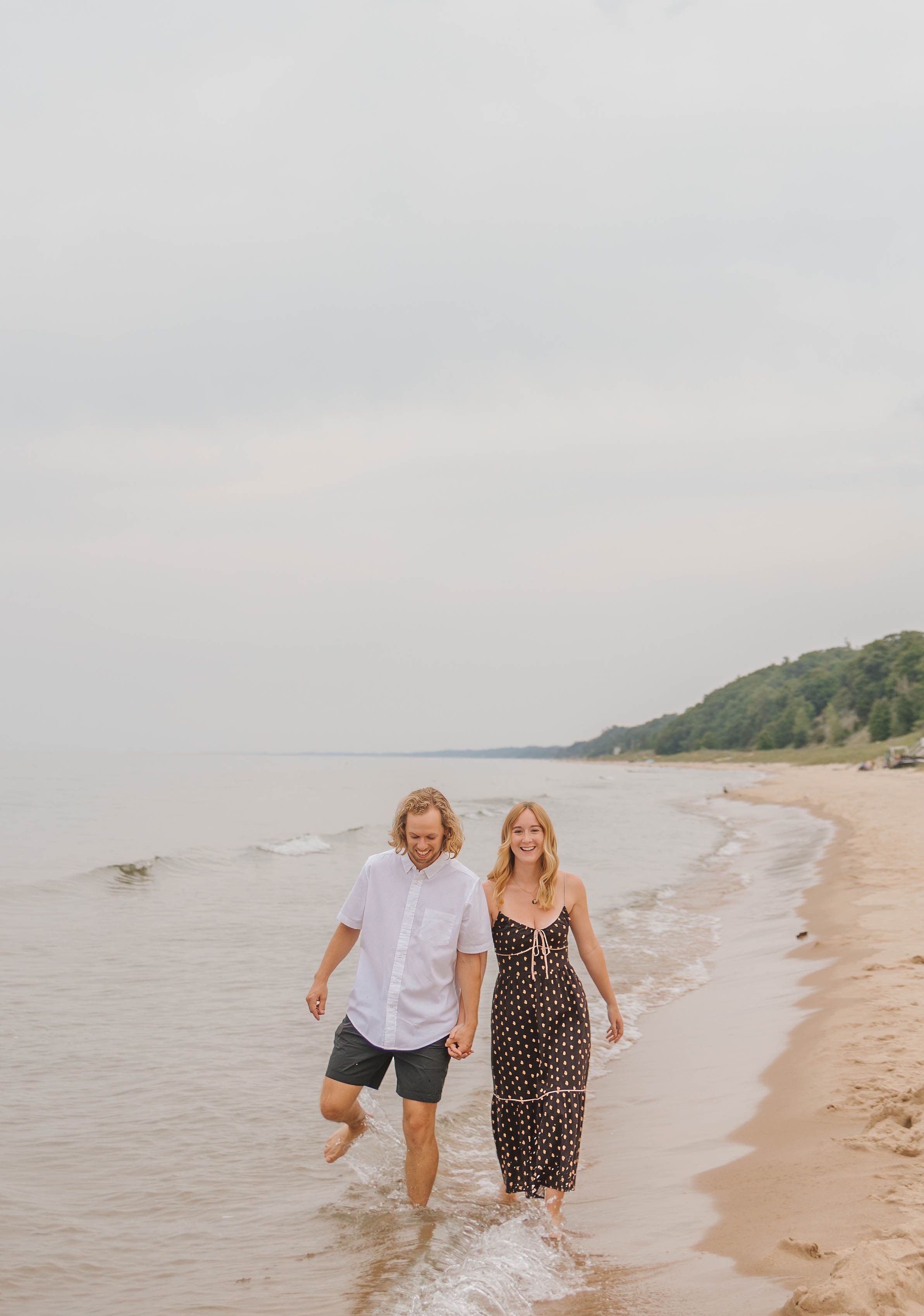 A couple walks hand-in-hand along a sandy beach shoreline, smiling, with calm water and green bluffs in the background.