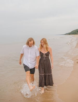 Couple walking hand-in-hand along a sandy beach shore, waves splashing at their feet on an overcast day.