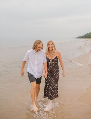 A couple walks barefoot along a sandy beach shoreline, smiling and holding hands, wearing casual summer outfits.