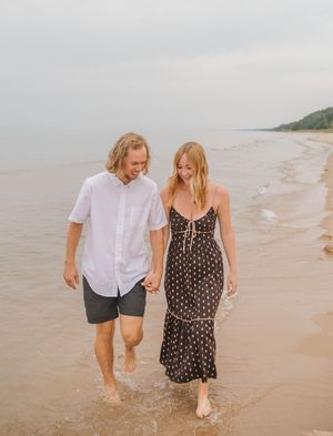 Couple walking hand in hand along a sandy beach shoreline, man in white shirt and woman in black floral dress.