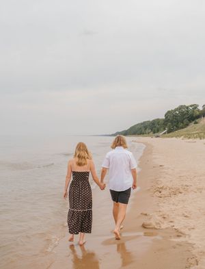 Couple holding hands walking along sandy beach shoreline in casual summer attire on overcast day.