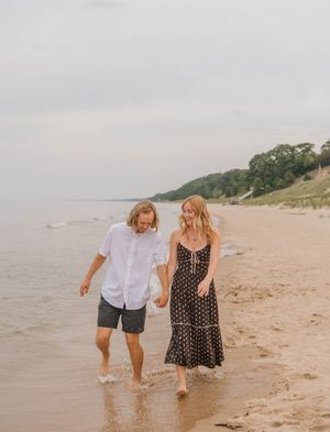 Couple walking hand-in-hand along a sandy beach shoreline, woman in floral dress and man in white shirt.