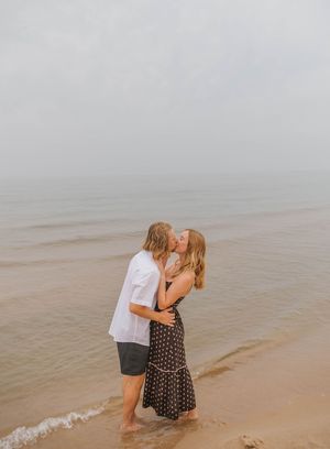 Couple sharing a romantic kiss on a misty beach shoreline, woman in floral dress and man in white shirt.