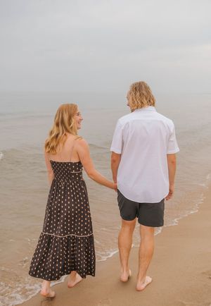 Couple holding hands walking barefoot on a sandy beach, woman in polka dot maxi dress, man in white shirt and dark shorts.