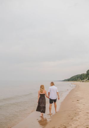 Couple strolling hand in hand along serene sandy beach with lush green bluffs in background.