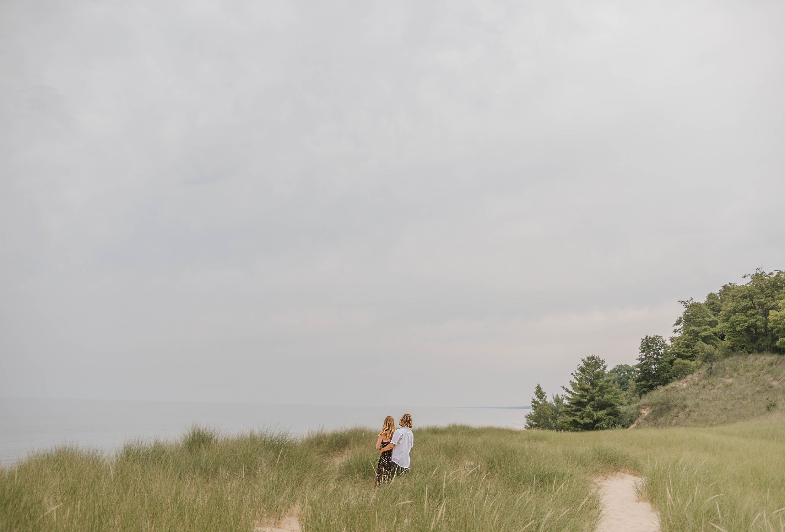 Two children walk along a sandy path through tall green dunes under a cloudy sky near the beach.