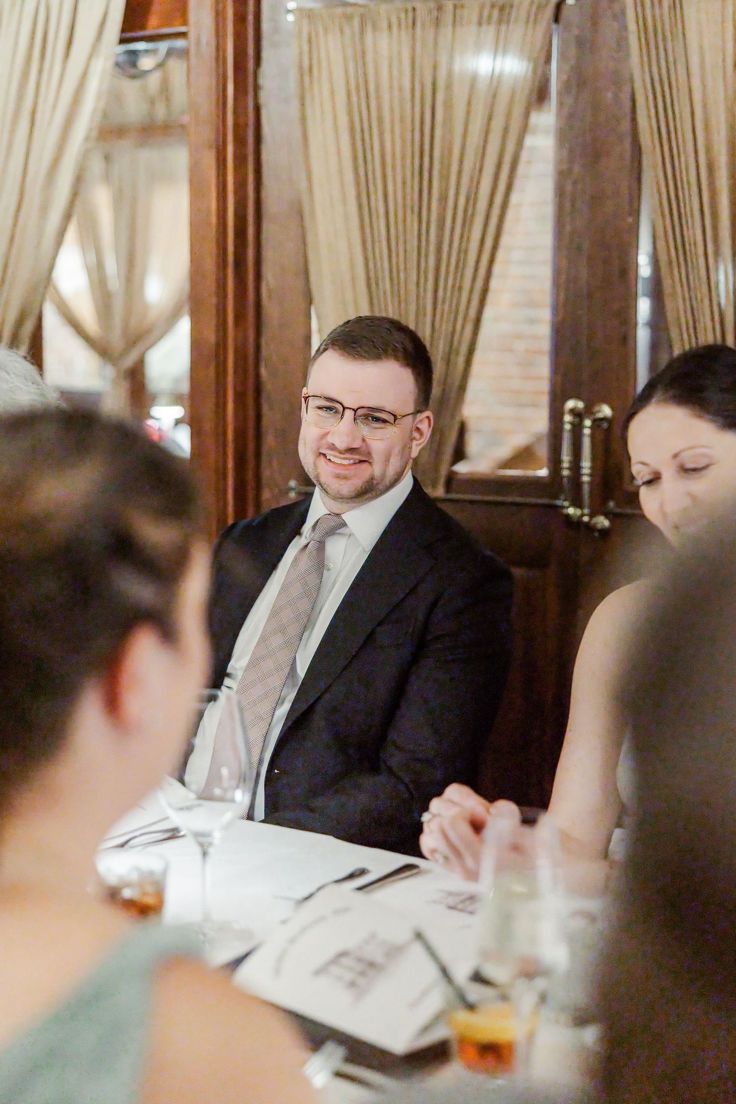 A smiling man in glasses and dark suit with tie sits at a formal dining table during an elegant restaurant gathering.