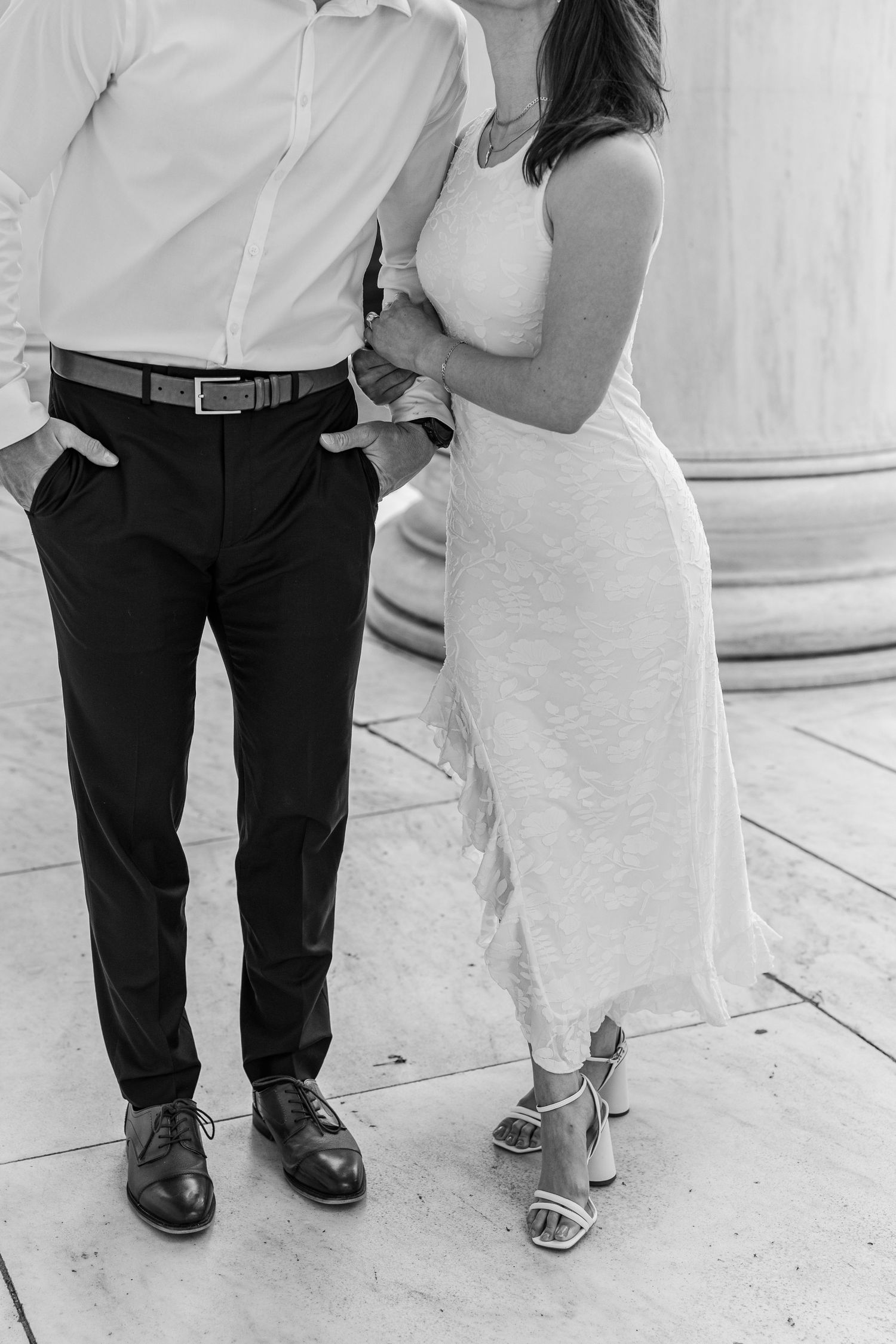 Black and white photo of a couple in formal attire, man in dress pants and woman in white dress, standing by a column.