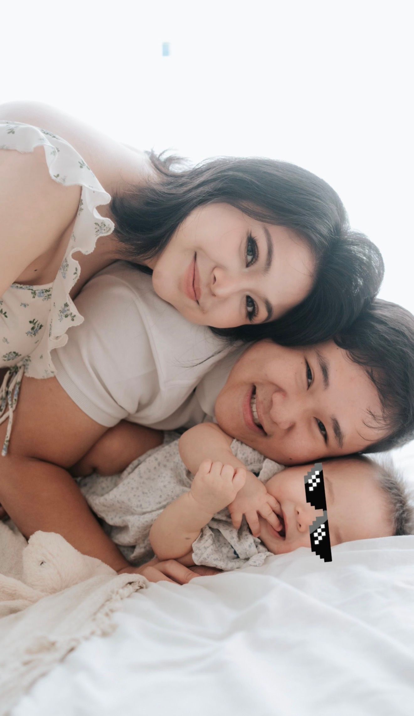 A smiling Asian family of three, mother, father, and baby with pixel sunglasses, lying together on a white bed.