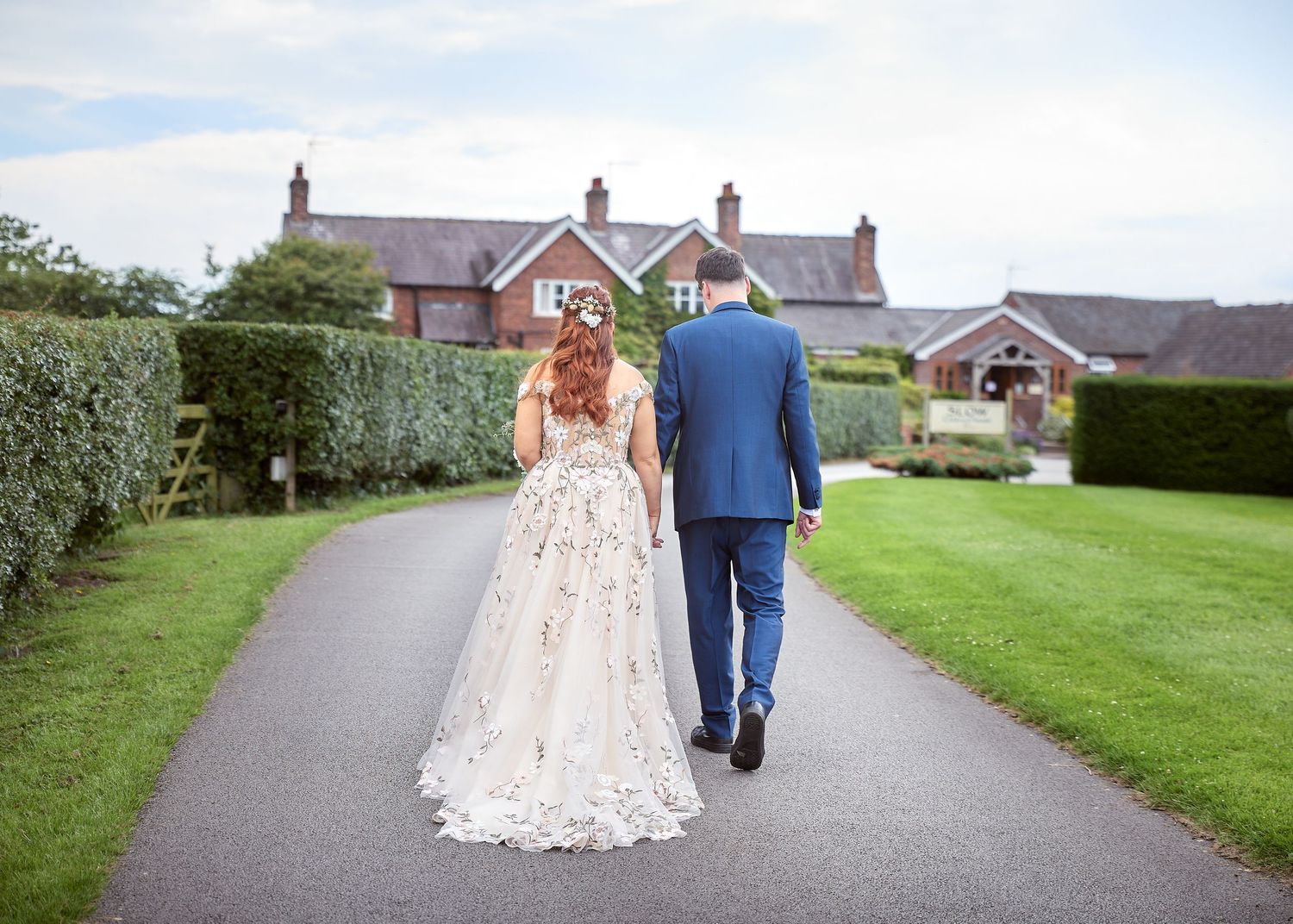 A bride and groom walk hand in hand along a garden path toward a brick country house on their wedding day.