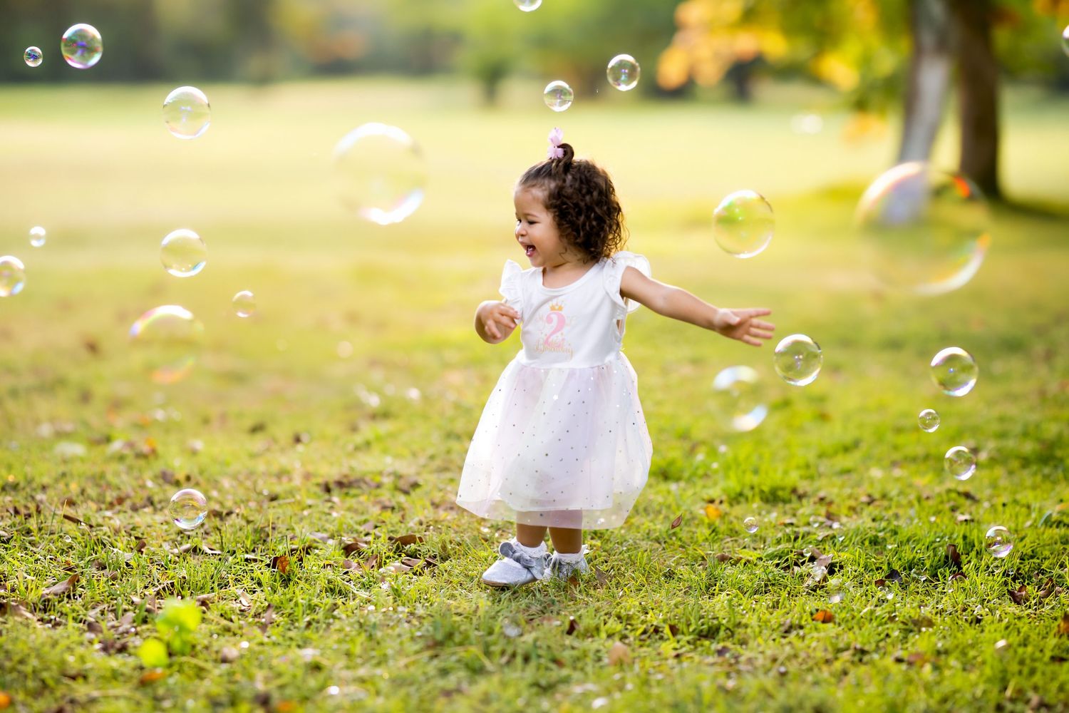 A joyful toddler girl in a white dress chasing soap bubbles in a sunny green park.