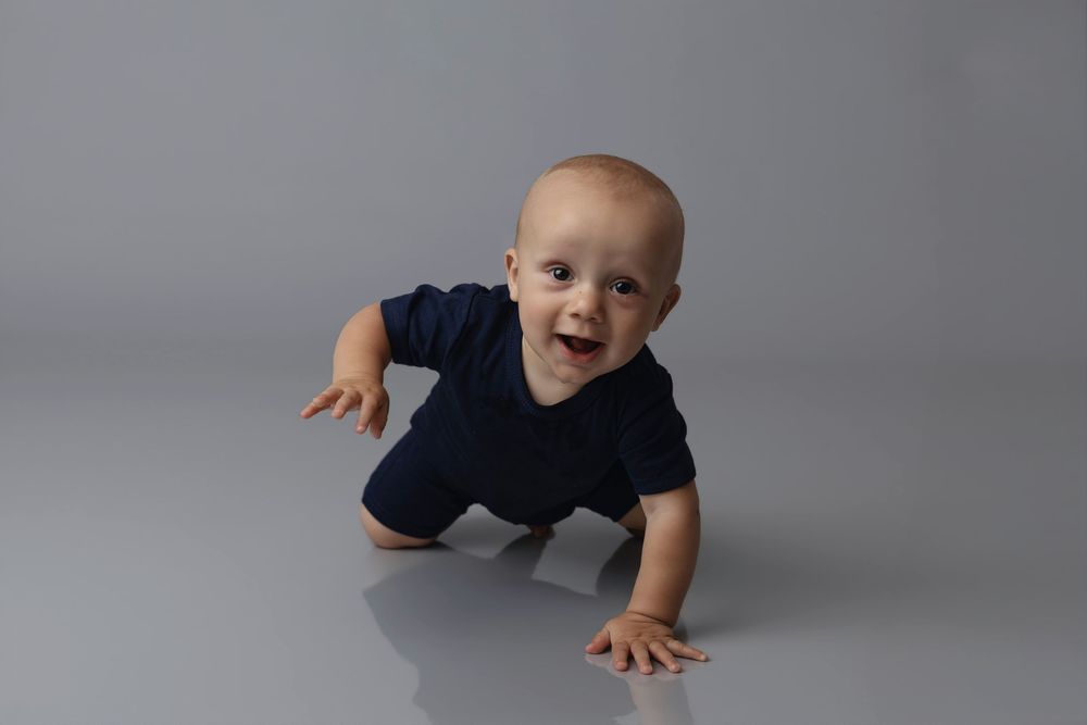 A happy baby in a navy blue onesie crawling on a gray studio floor, smiling brightly at the camera.