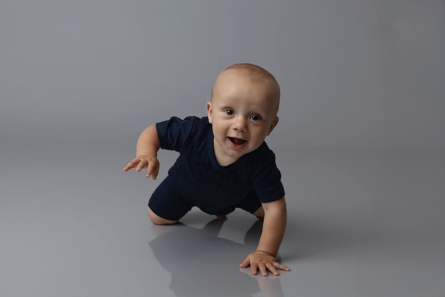 A happy baby in a navy blue onesie crawling on a gray studio floor, smiling brightly at the camera.