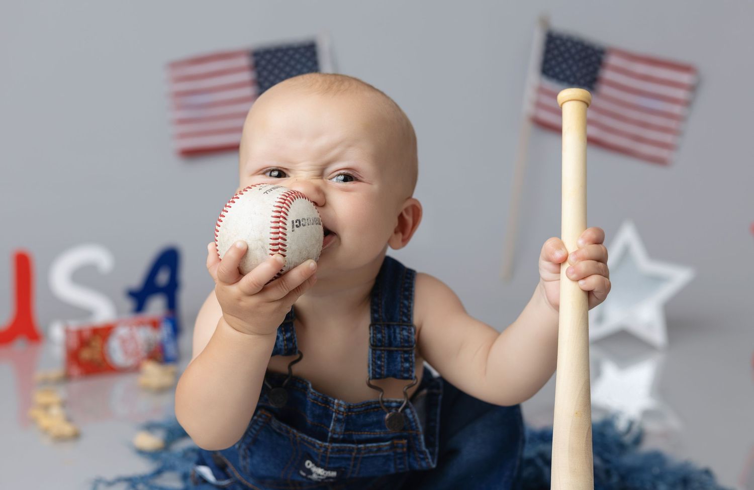Baby in denim overalls holding a baseball and bat with American flags for a patriotic USA photo shoot.
