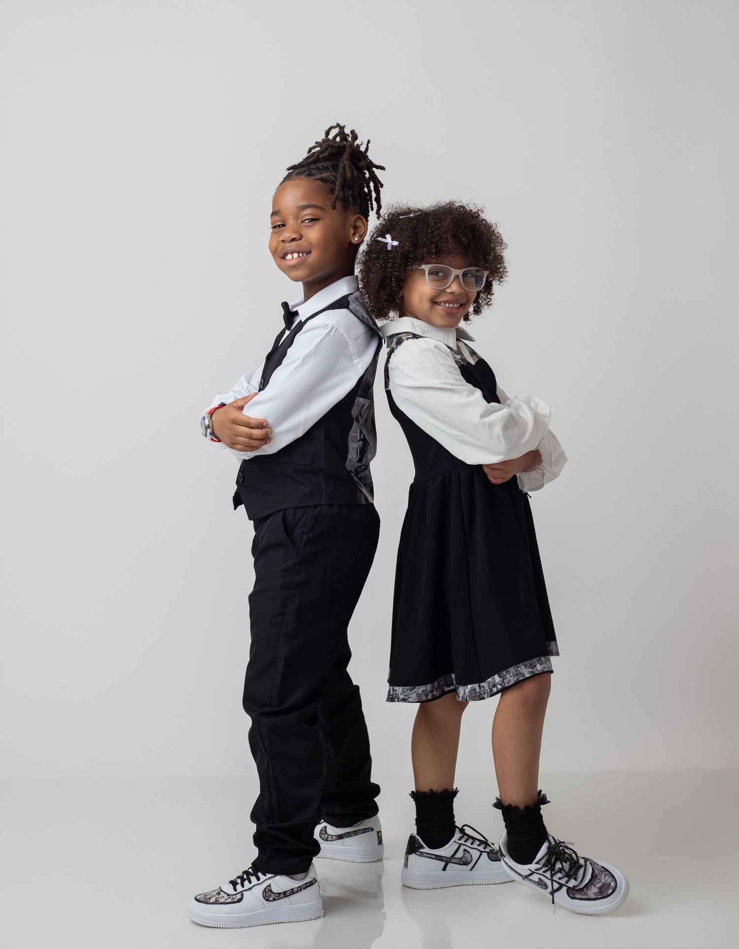 Two stylish children in black and white outfits pose back-to-back wearing Nike sneakers against a gray background.