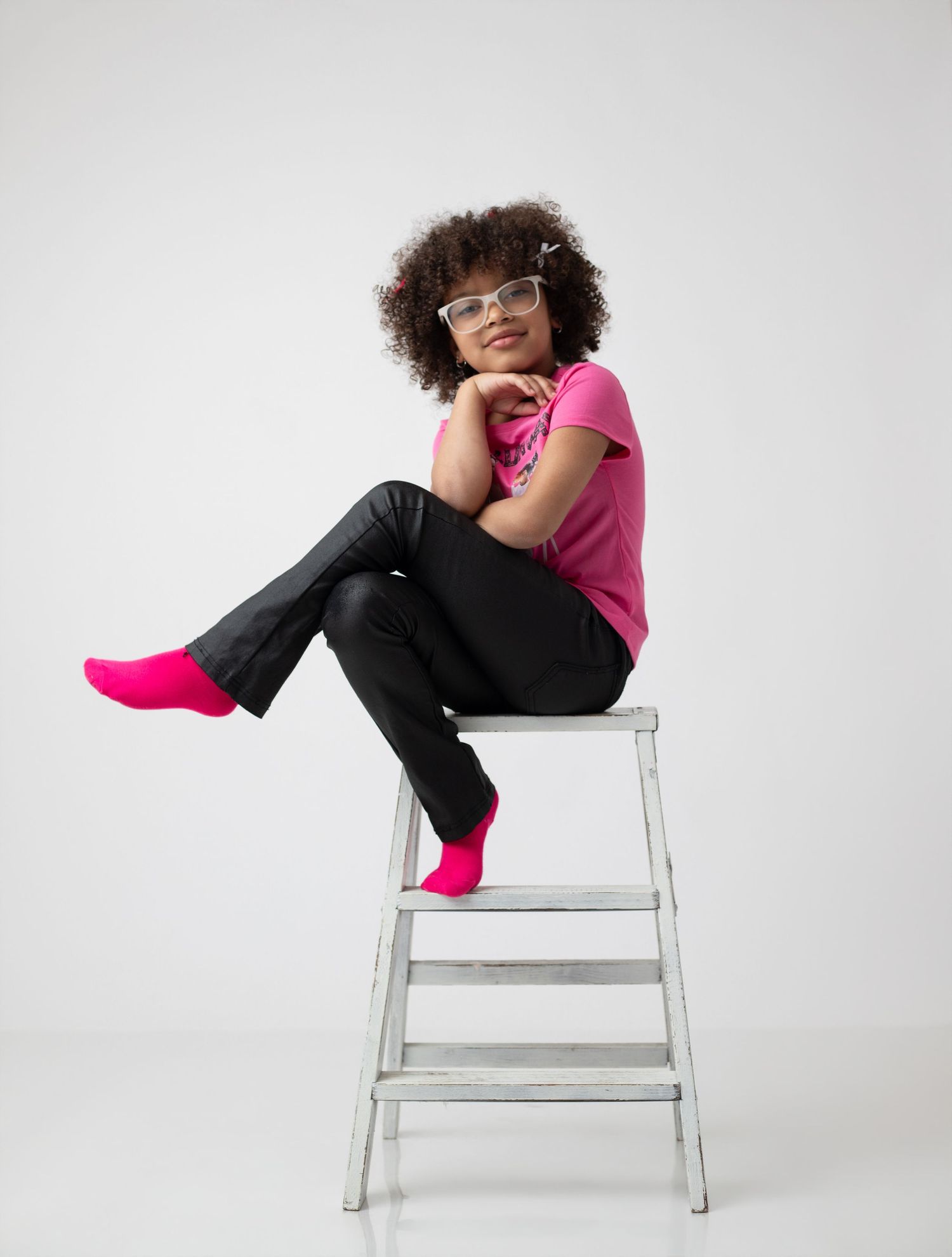 Young woman with curly hair sits on a metal step stool, wearing pink tee, black pants, and bright pink socks.