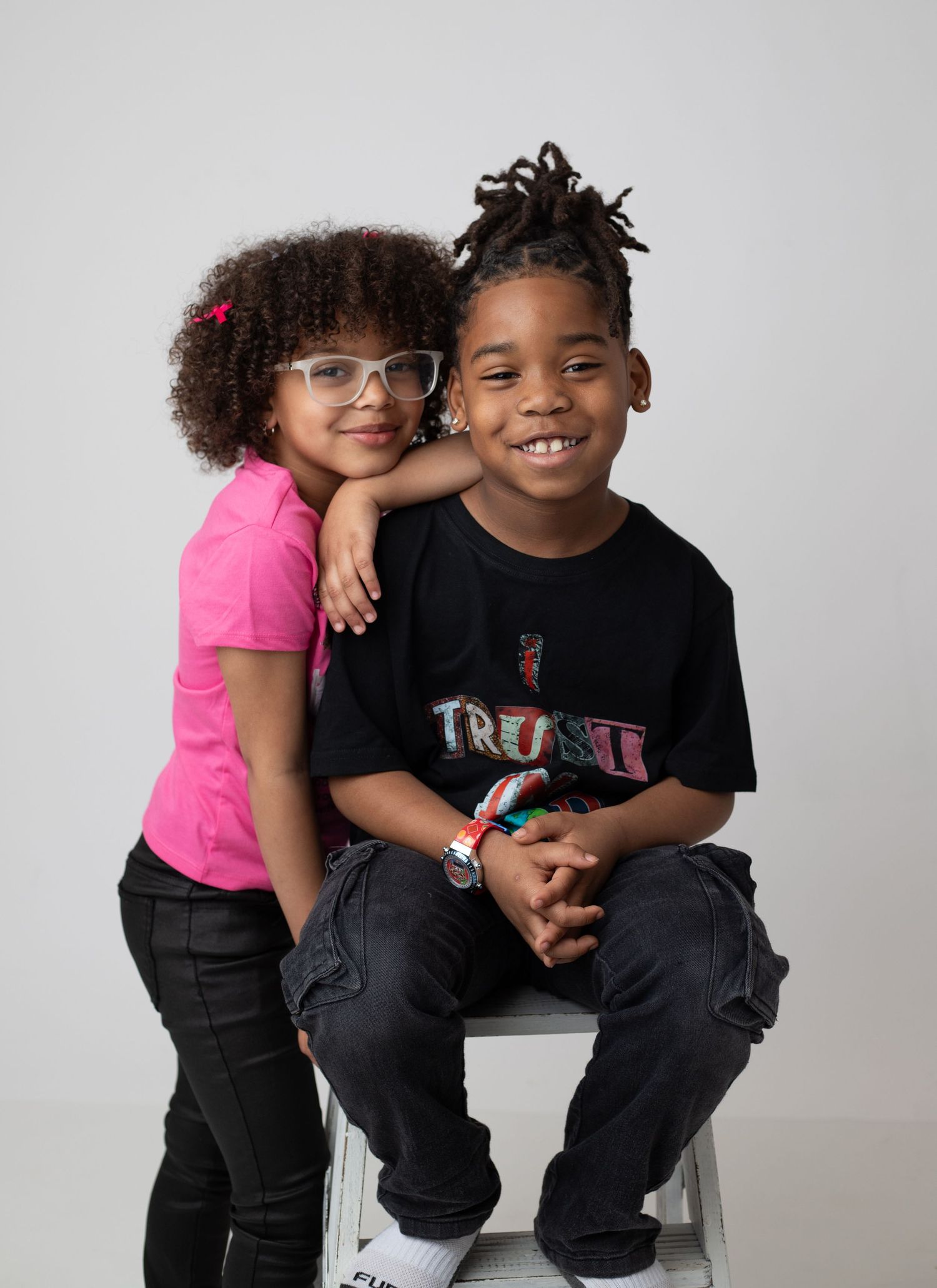Two smiling children pose together, a girl in pink shirt with glasses and a boy in black tee sitting on a white box.