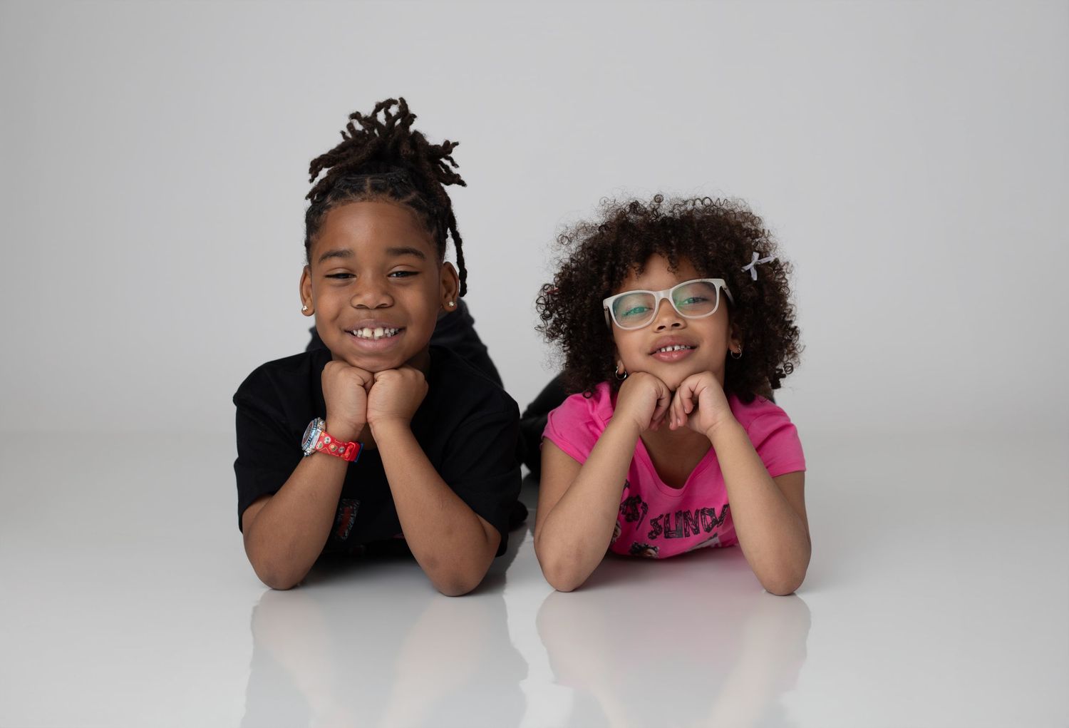 Two young children lying on floor smiling, girl with glasses in pink shirt and boy in black shirt, studio portrait.