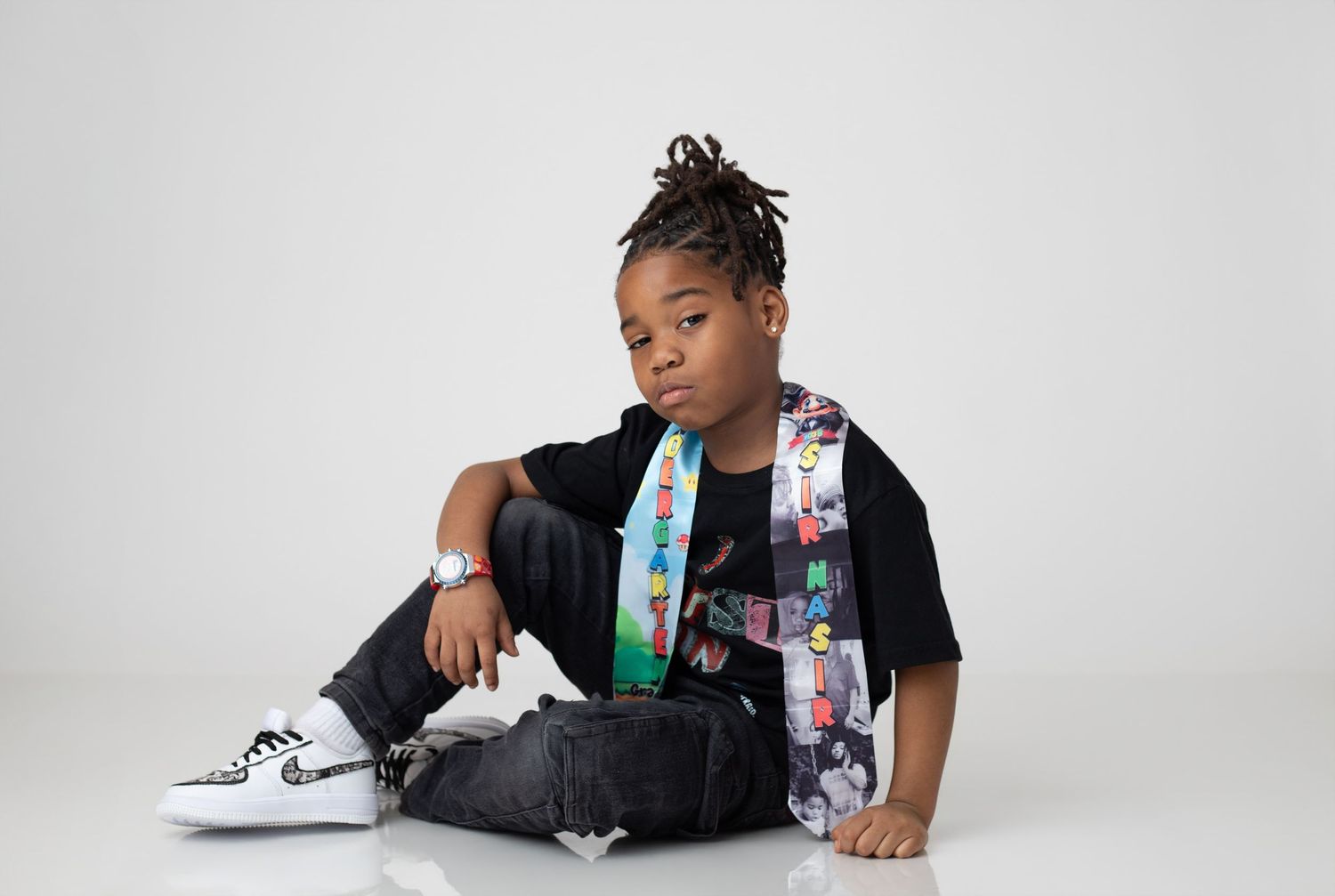 Young child with braids wearing black tee, jeans, white Nike sneakers, and colorful sash, posing on white background.