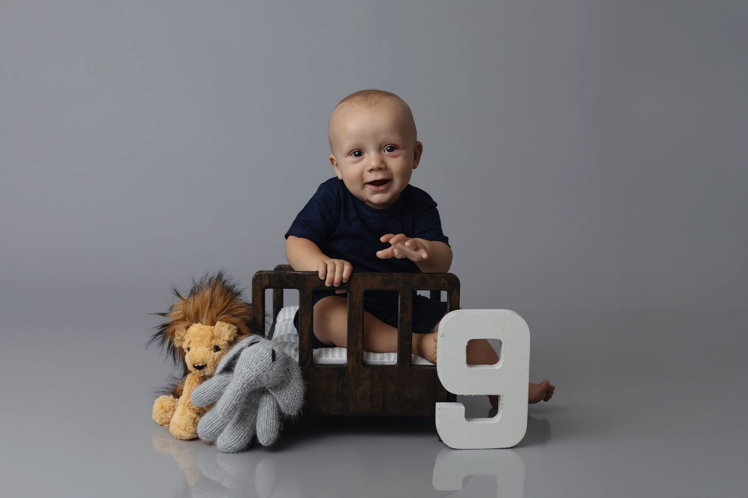 Smiling baby sitting in wooden crate with stuffed lion and elephant toys beside number 9, studio portrait photo.