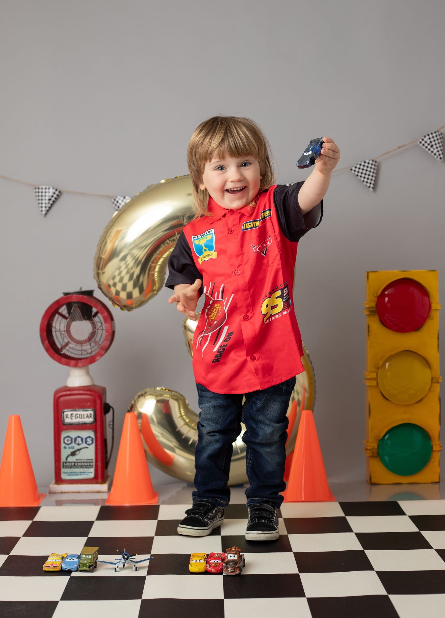 Toddler boy in racing outfit holds toy car on checkered floor with traffic light, cones, and number 2 balloon for birthday.