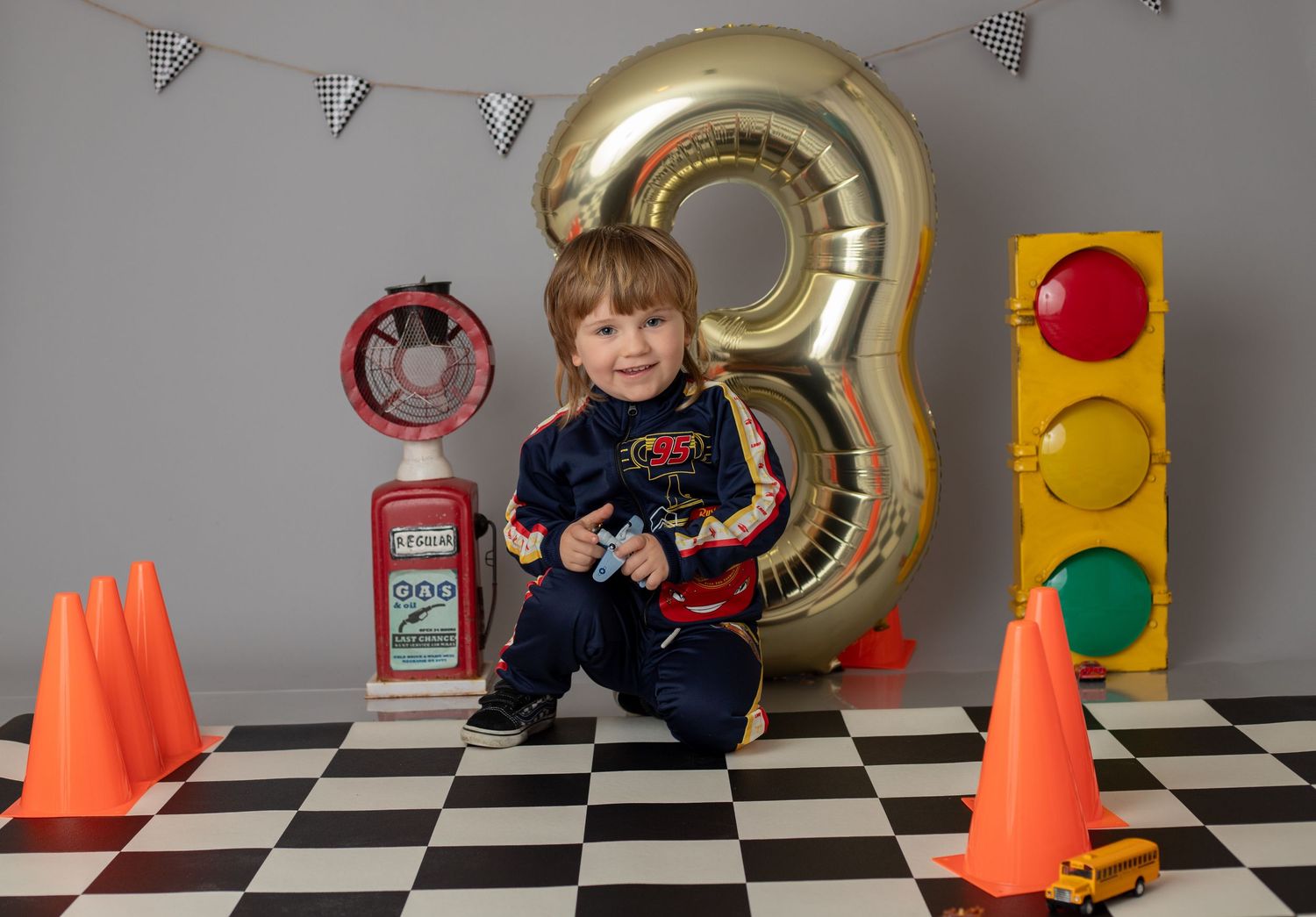 Boy in racing suit sits by a large gold number 8 balloon with traffic lights and cones for a race-themed 8th birthday.