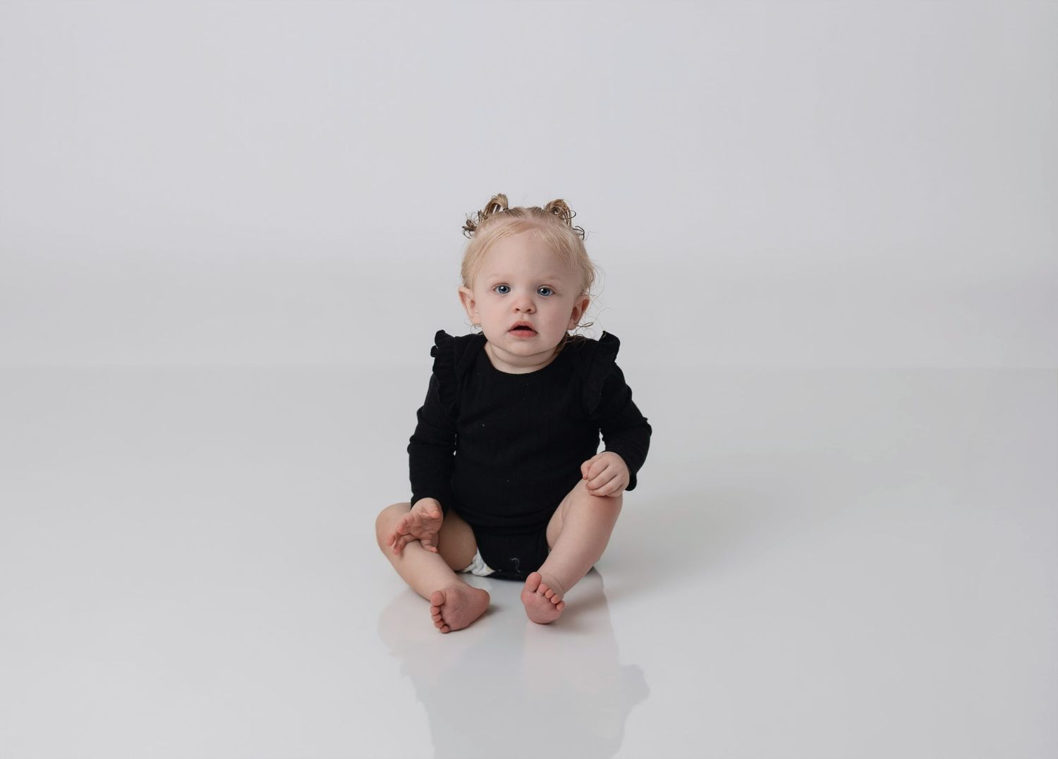 Baby girl in black ruffle bodysuit sitting on white background with small hair clips.