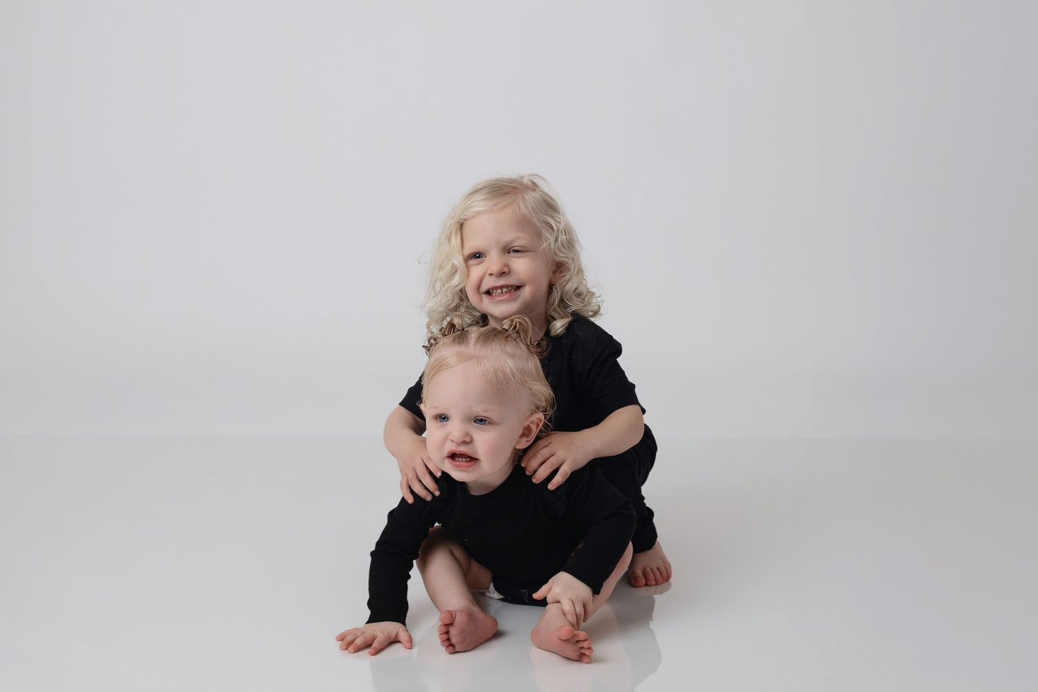 Two blonde toddlers in black outfits pose together on white background, older child hugging younger sibling.